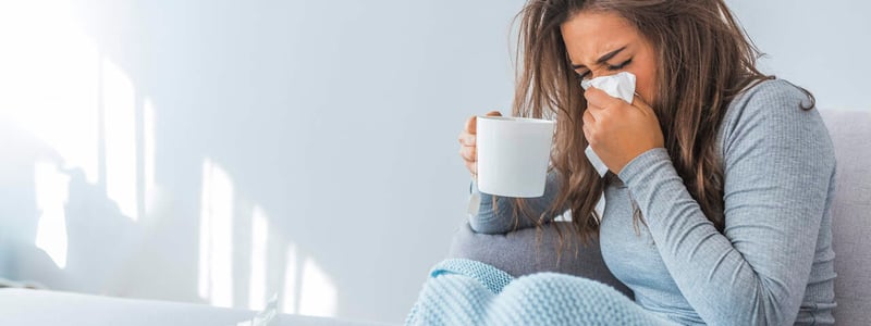 Eine erkältete Frau hält eine Teetasse in der einen Hand und putzt sich mit der andere Hand die Nase.