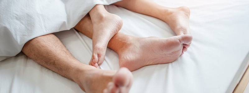 In einem weiß bezogenen Bett gucken die Füße von einem Mann und einer Frau unten aus der Bettdecke.