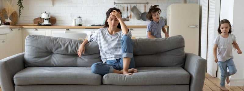 Eine Frau sitzt genervt auf dem Sofa, während Kinder um sie herumtoben.