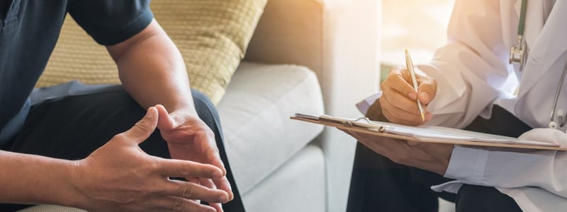 Ein Patient sitzt auf der Couch, ihm gegenüber sitzt ein Arzt, der sich Notizen macht.