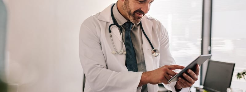 Arzt sitzt auf der Ecke eines Schreibtischs und nutzt ein Tablet.