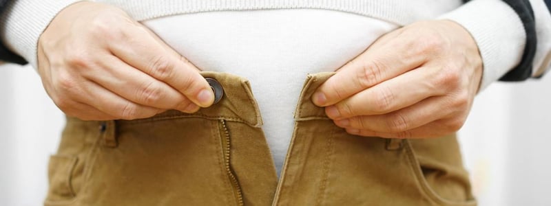 Person bekommt Knopf an der braunen Hose über dem Bauch nicht geschlossen.