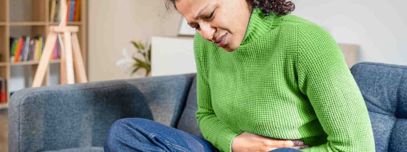 Frau sitzt zusammengekrümmt auf dem Sofa und hält sich den schmerzenden Bauch.