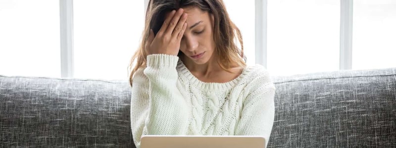 Eine Frau sitzt mit Laptop auf dem Sofa und hat Kopfschmerzen.