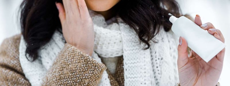 Eine junge Frau trägt Hautpflege auf ihr Gesicht auf. Sie trägt winterliche Kleidung.