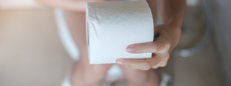 Eine Frau sitzt auf der Toilette und hält eine Rolle Klopapier in der Hand.
