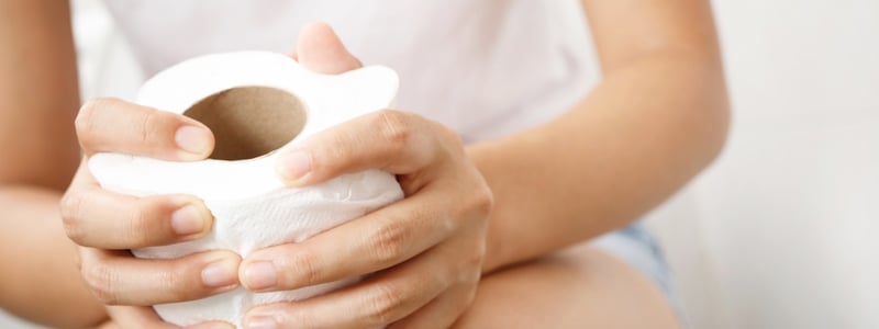 Frau sitzt auf der Toilette und hält eine Rolle Klopapier in den Händen.