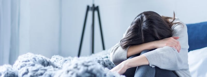 Junge Frau in grauer Kleidung sitzt zusammengerollt auf dem Bett zu Hause. 
