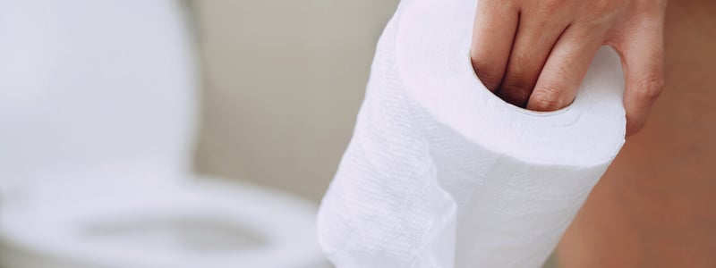 Eine Frauenhand hält eine Rolle Klopapier, im Hintergrund ist eine Toilette zu sehen.