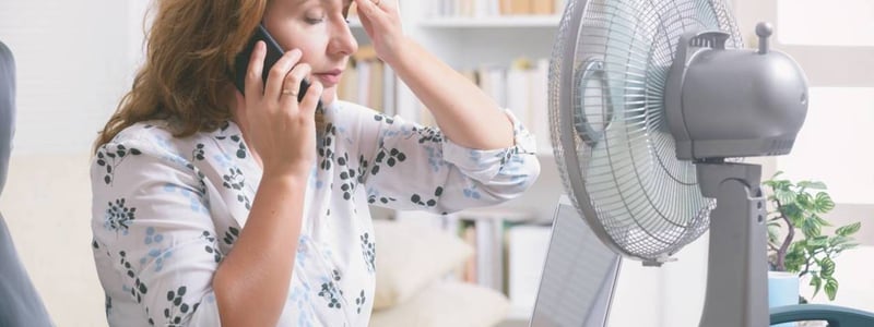 Frau am Telefon sitzt vor dem Ventilator und tupft sich die Stirn mit einem Tuch.