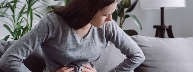 Eine junge Frau sitzt auf dem Sofa und hält sich vor Schmerzen den Bauch.