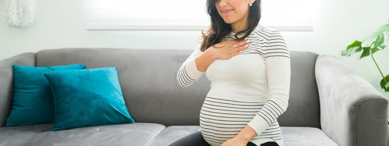 Schwangere Frau sitzt auf dem Sofa. Sie hält sich die Brust und hat Schmerzen vom Sodbrennen.