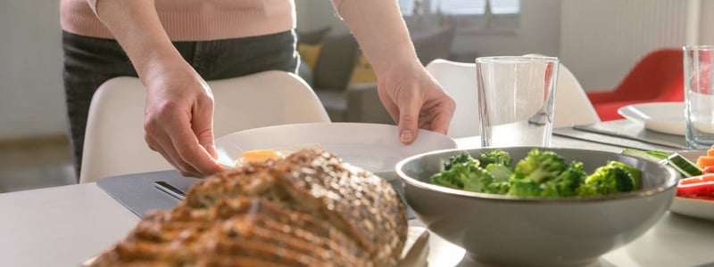 Im Bild ist ein gedeckter Tisch zu sehen, darauf aufgeschnittenes Brot und Broccoli. Eine Frau stellt einen Teller auf den Tisch.