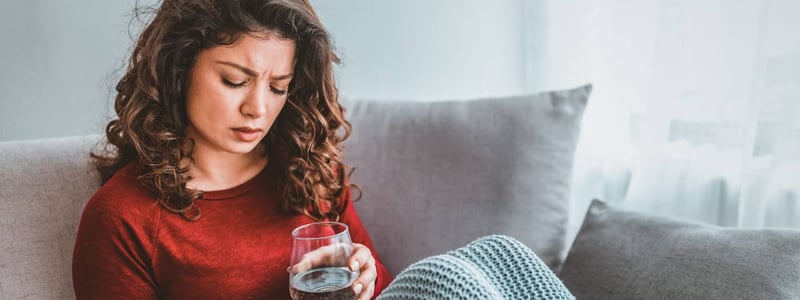 Eine Frau sitzt auf dem Sofa und sieht beunruhigt aus. Sie hält ein Glas Wasser und eine Tablette in den Händen.