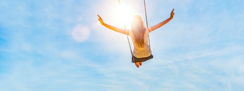 Frau schaukelt vor blauem Himmel und Sonne, sie streckt die Arme zur Seite.