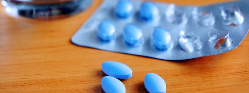 Blaue Tabletten in einem Blister liegen auf einem Holztisch, im Hintergrund steht ein Glas Wasser.