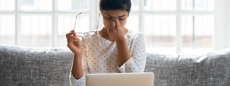 Eine junge Frau sitzt auf der Couch, vor ihr steht ein Laptop. Sie hat ihre Brille abgenommen, schließt die Augen und wirkt gestresst.