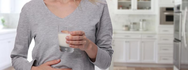 Laktoseintoleranz: Frau hält Glas mit Milch in der einen Hand, mit der anderen Hand hält sie ihren schmerzenden Bauch.