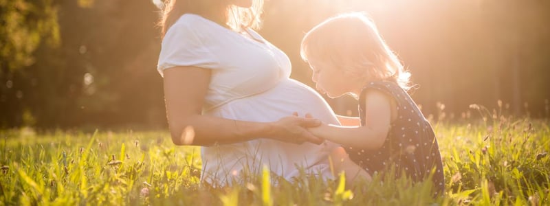 Schwangere Frau sitzt mit Mädchen auf einer Wiese in der Sonne.
