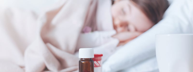 Schlaftabletten stehen auf einem Nachttisch, im Hintergrund schläft eine Frau im Bett.