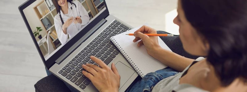 Eine Frau sitzt am Laptop und macht sich während der Online-Sprechstunde mit einer Ärztin Notizen.