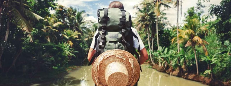 Frau macht einen Abenteuerurlaub. Sie steht mit Rucksack und Strohhut an einem Fluss.