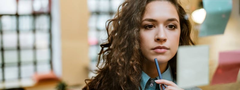 Eine junge Frau schaut konzentriert mit einem Stift am Kinn auf eine Glaswand mit Post-Its.