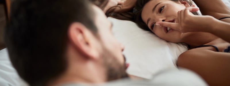 Mann und Frau liegen im Bett. Die Frau hält ihren Zeigefinger an ihre Lippen.