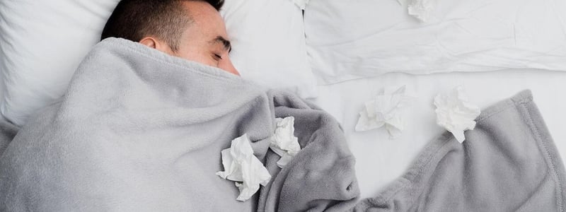 Ein Mann liegt im Bett. Er ist bis zur Nase zugedeckt und es liegen viele benutzte Taschentücher um ihn herum.